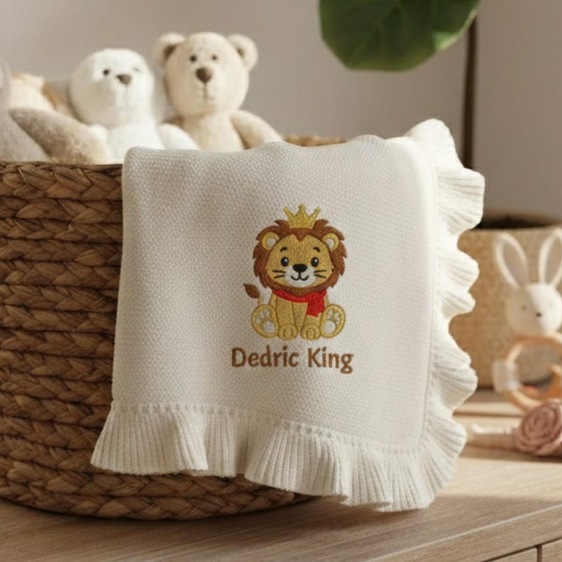 May include: Cream-colored baby blanket with a ruffled edge. The blanket features an embroidered lion wearing a crown and a red scarf, with the text "Dedric King" below. The blanket is folded over a wicker basket, with stuffed animals in th