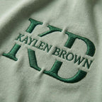 May include: Close-up of a light green fabric with the embroidered text "KAYLEN BROWN" in a dark green color. The letters are stylized and bold, creating a textured appearance against the smooth background.