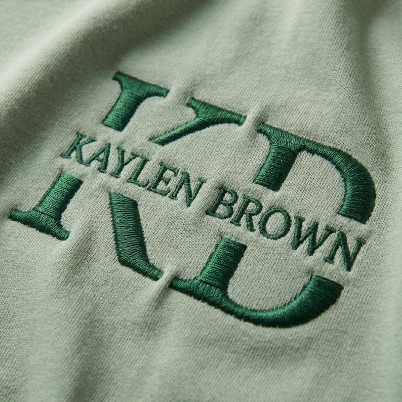 May include: Close-up of a light green fabric with the embroidered text "KAYLEN BROWN" in a dark green color. The letters are stylized and bold, creating a textured appearance against the smooth background.