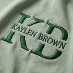 May include: Close-up of a light green fabric with the embroidered text "KAYLEN BROWN" in a dark green color. The letters are stylized and bold, creating a textured effect against the smooth background.