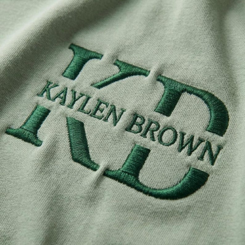 May include: Close-up of a light green fabric with the embroidered text "KAYLEN BROWN" in a dark green color. The letters are stylized and bold, creating a textured effect against the smooth background.