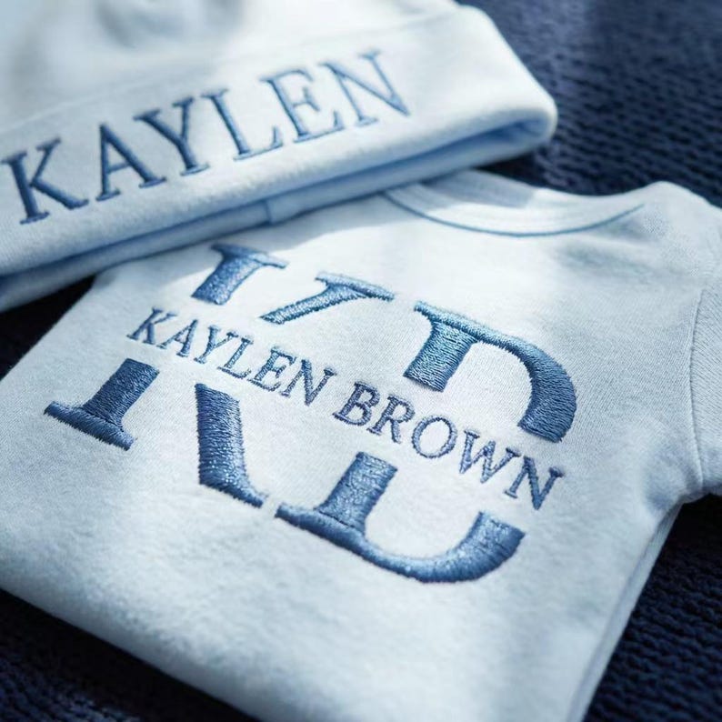 May include: A light blue baby hat and onesie set. The hat has the name "KAYLEN" embroidered in blue. The onesie features the name "KAYLEN BROWN" embroidered in a stylized font. The set is made of soft material.