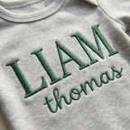 May include: A gray baby onesie with the name "LIAM" embroidered in dark green capital letters above the name "thomas" in cursive. The onesie has a round neckline and short sleeves.