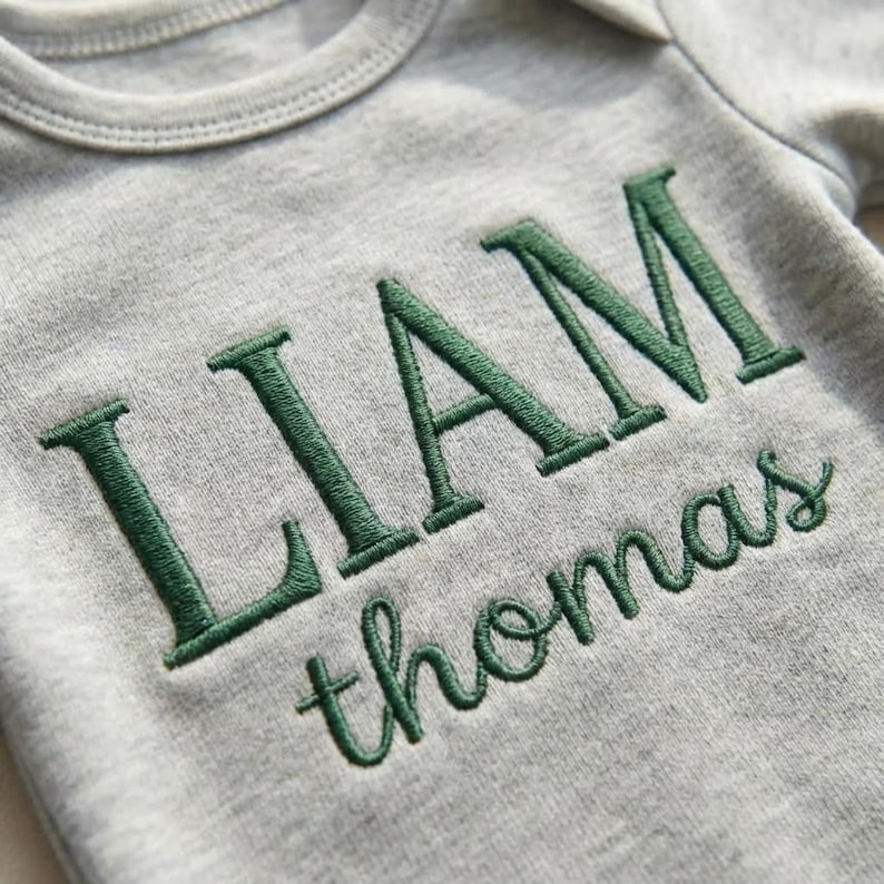 May include: A gray baby onesie with the name "LIAM" embroidered in dark green capital letters above the name "thomas" in cursive. The onesie has a round neckline and short sleeves.