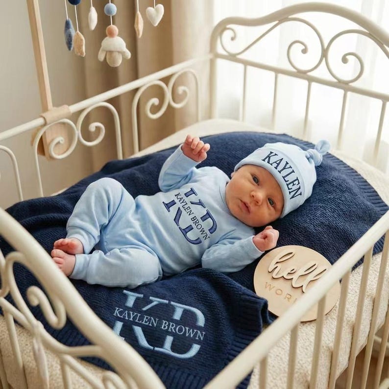 May include: A baby in a light blue outfit and matching hat, lying on a navy blue blanket inside a white crib. The outfit and blanket are personalized with the name "Kaylen Brown". A wooden sign that says "hello world" is also in the crib.