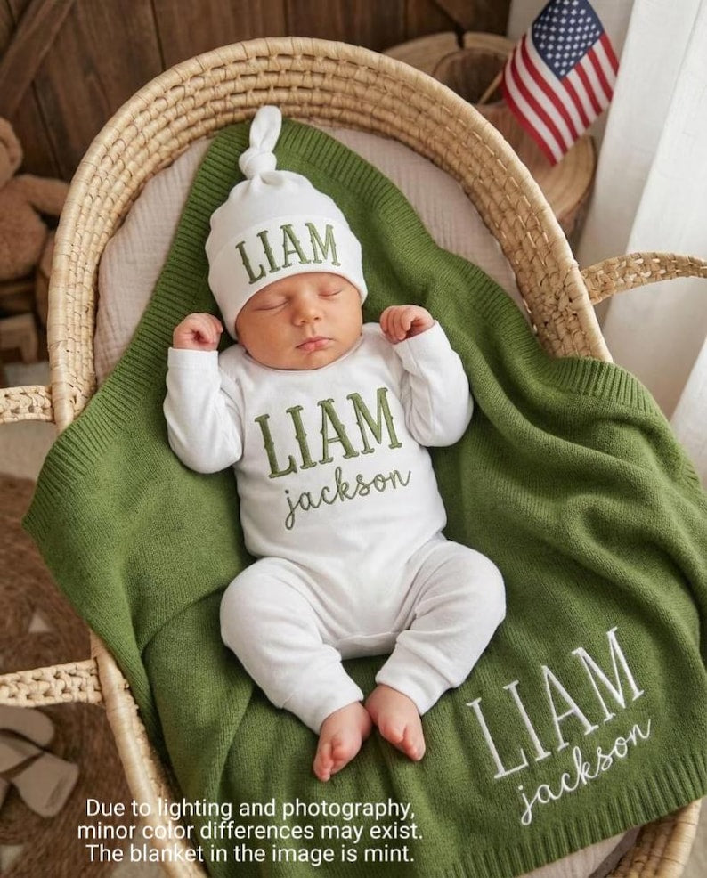 May include: A newborn baby wearing a white hat and onesie, both embroidered with the name "LIAM" in green. The baby is lying on a mint green blanket, also embroidered with "LIAM Jackson", inside a woven basket. An American flag is in the b