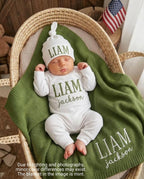 May include: A newborn baby wearing a white hat and onesie, both embroidered with the name "LIAM" in green. The baby is lying on a mint green blanket, also embroidered with "LIAM Jackson", inside a woven basket. An American flag is in the b