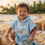 May include: A light blue hooded beach towel with the name "Elijah" embroidered on the front. The towel features a waffle weave texture and a pocket with crab, bucket, and sandcastle designs. The child is holding a sand bucket and a small s
