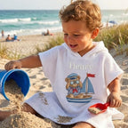 May include: A white hooded waffle-weave beach towel with the name "Henry" embroidered on it. The towel features a nautical-themed graphic of a teddy bear in a sailor outfit on a sailboat. A child is playing with sand on the beach.