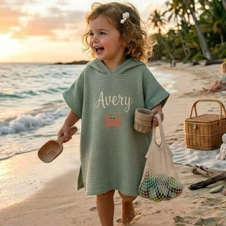 May include: A light green hooded waffle-weave beach cover-up with the name "Avery" and a crab graphic. The child is holding a wooden scoop, a small cup, and a mesh bag with colorful items. A wicker basket sits on the beach.