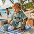 May include: A light green waffle-weave hooded beach towel with the name "Henry" embroidered in black and an anchor design. A blue bucket with the initials "HJC" sits on a blue and white striped towel. A wicker basket is also visible.