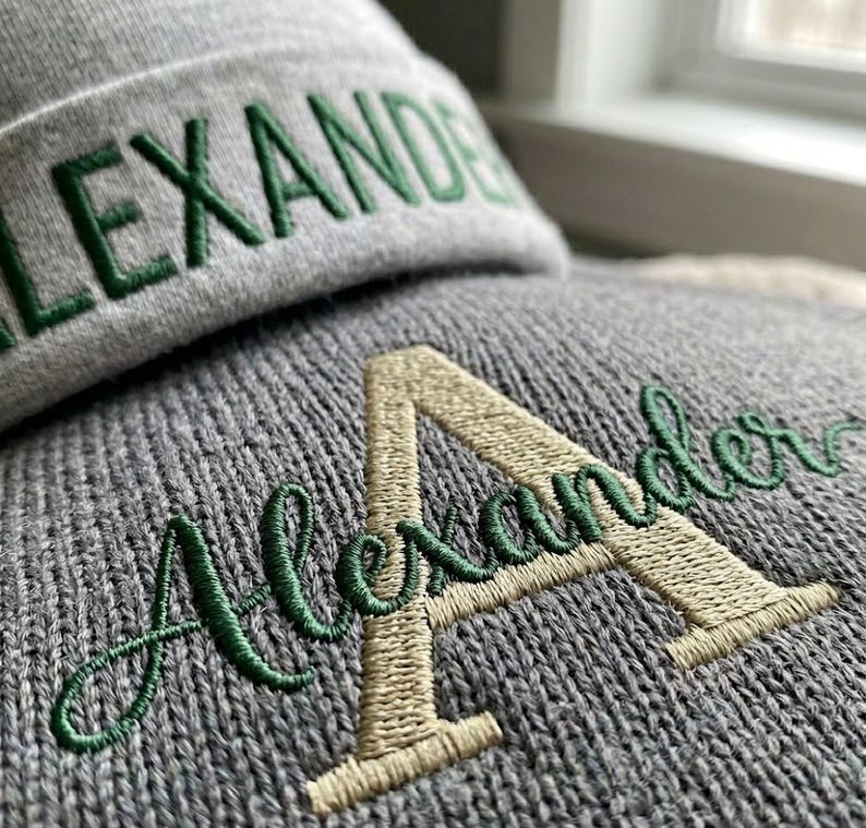 May include: A grey knit beanie featuring the embroidered name ALEXANDER in dark green block letters on the cuff. Below the cuff, a grey knit fabric displays a large, light-colored embroidered letter A with the name Alexander in green scrip