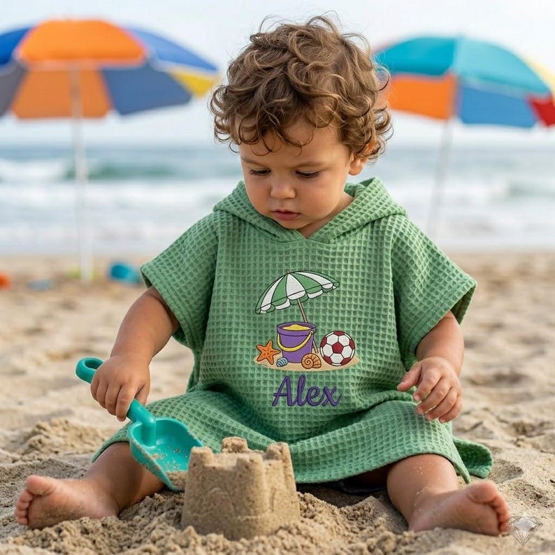 May include: A green waffle-weave hooded beach poncho with a beach-themed graphic, including a purple bucket, soccer ball, and umbrella. The name "Alex" is embroidered on the front. A child is playing with a sandcastle and a blue shovel on 