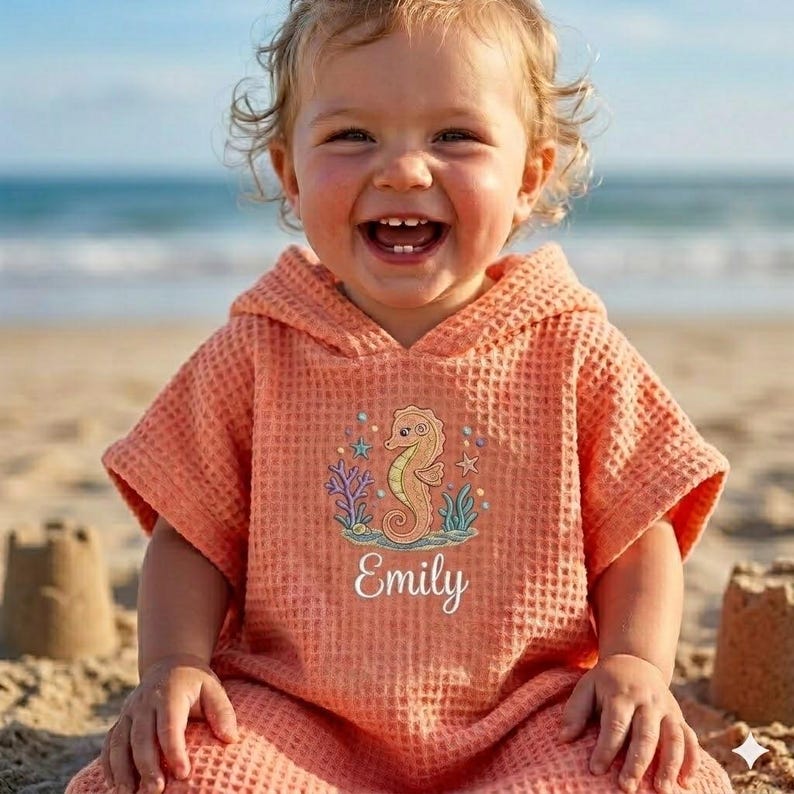 May include: A coral-colored hooded beach towel with a seahorse design and the name "Emily" embroidered on the front. The towel has a waffle weave texture and is designed for children. The background shows a beach and ocean.