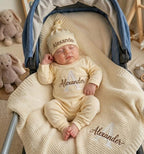 May include: A cream-colored long-sleeve onesie and matching beanie set featuring the name Alexander embroidered in brown thread. The onesie displays a large letter A with the name Alexander, and a matching cream knit blanket features the s