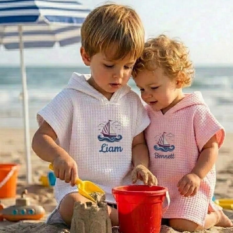 May include: Two children wearing hooded beach towels on a sandy beach. One towel is white with a sailboat graphic and the name "Liam" in blue. The other towel is pink with a sailboat graphic and the name "Bennett" in blue. They are playing