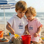 May include: Two children wearing hooded beach towels on a sandy beach. One towel is white with a sailboat graphic and the name "Liam" in blue. The other towel is pink with a sailboat graphic and the name "Bennett" in blue. They are playing
