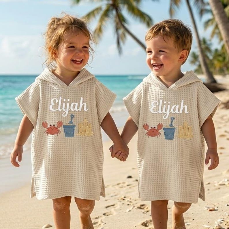 May include: Two children wearing beige hooded beach ponchos with the name "Elijah" printed on the front. Each poncho features a crab, bucket, and sandcastle design. The children are holding hands on a sandy beach with palm trees in the bac