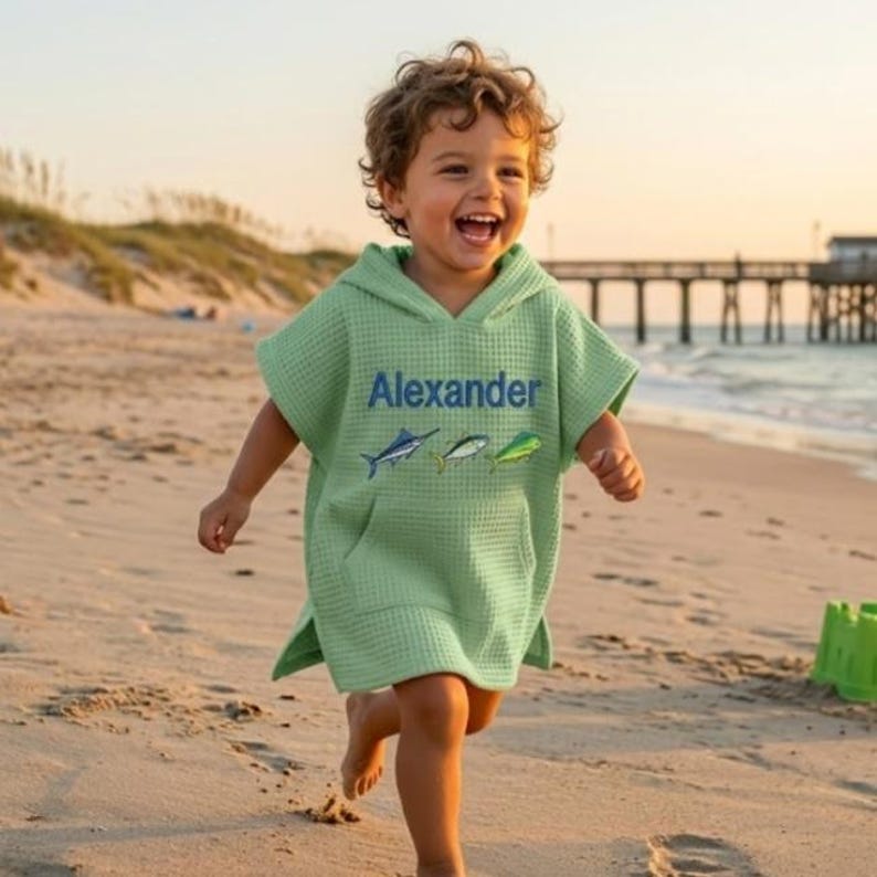 May include: A light green hooded beach towel with the name "Alexander" embroidered in blue, along with three fish illustrations. The towel is worn by a child running on a sandy beach near the ocean.