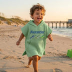 May include: A light green hooded beach towel with the name "Alexander" embroidered in blue, along with three fish illustrations. The towel is worn by a child running on a sandy beach near the ocean.