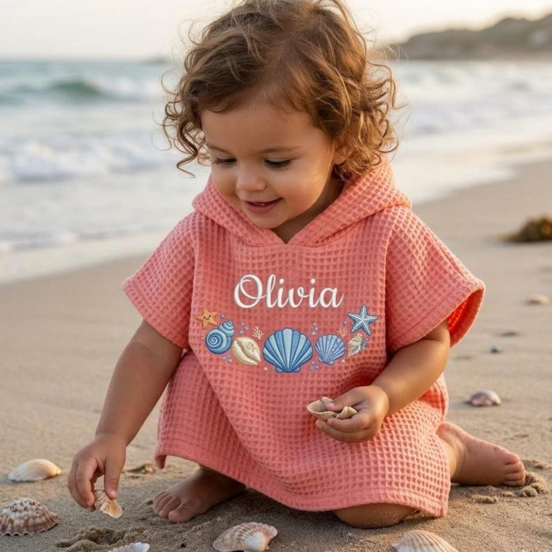 May include: A coral-colored hooded beach towel with the name "Olivia" embroidered in white script. The towel features embroidered seashells and a starfish design. The towel is made of a waffle-weave fabric.