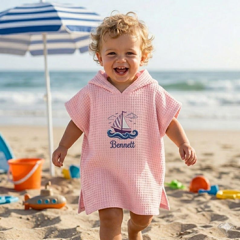 May include: A pink hooded beach towel with a sailboat design and the name "Bennett" embroidered on the front. The towel is made of a waffle-weave fabric and is being worn by a child on a sandy beach.