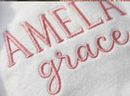 May include: Close-up of white fabric embroidered with the name "AMELIA" in capital letters and the word "grace" in cursive, both in a soft pink thread. The embroidery is detailed and the fabric appears to be a soft material.