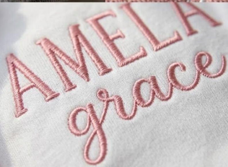May include: Close-up of white fabric embroidered with the name "AMELIA" in capital letters and the word "grace" in cursive, both in a soft pink thread. The embroidery is detailed and the fabric appears to be a soft material.