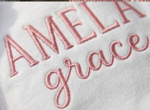 May include: Close-up of white fabric embroidered with the name "AMELIA" in capital letters and the word "grace" in cursive, both in a soft pink thread. The embroidery is detailed and the fabric appears to be a soft material.