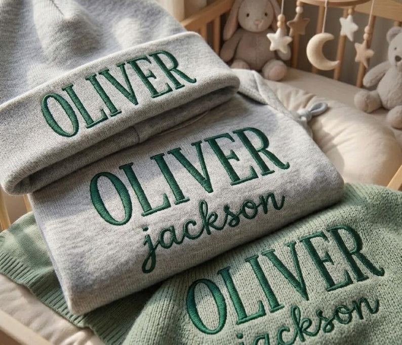 May include: A gray beanie, sweater, and blanket set with the name "OLIVER" embroidered in green. The sweater and blanket also feature the name "jackson". The items are displayed on a crib with stuffed animals and a mobile in the background