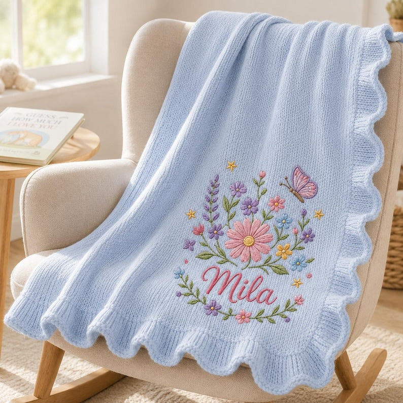 May include: A light blue knitted baby blanket with a scalloped edge. The blanket features an embroidered floral arrangement in pink, purple, yellow, and blue, a small pink butterfly, and the name Mila stitched in pink cursive script letter
