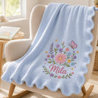 May include: A light blue knitted baby blanket with a scalloped edge. The blanket features an embroidered floral arrangement in pink, purple, yellow, and blue, a small pink butterfly, and the name Mila stitched in pink cursive script letter