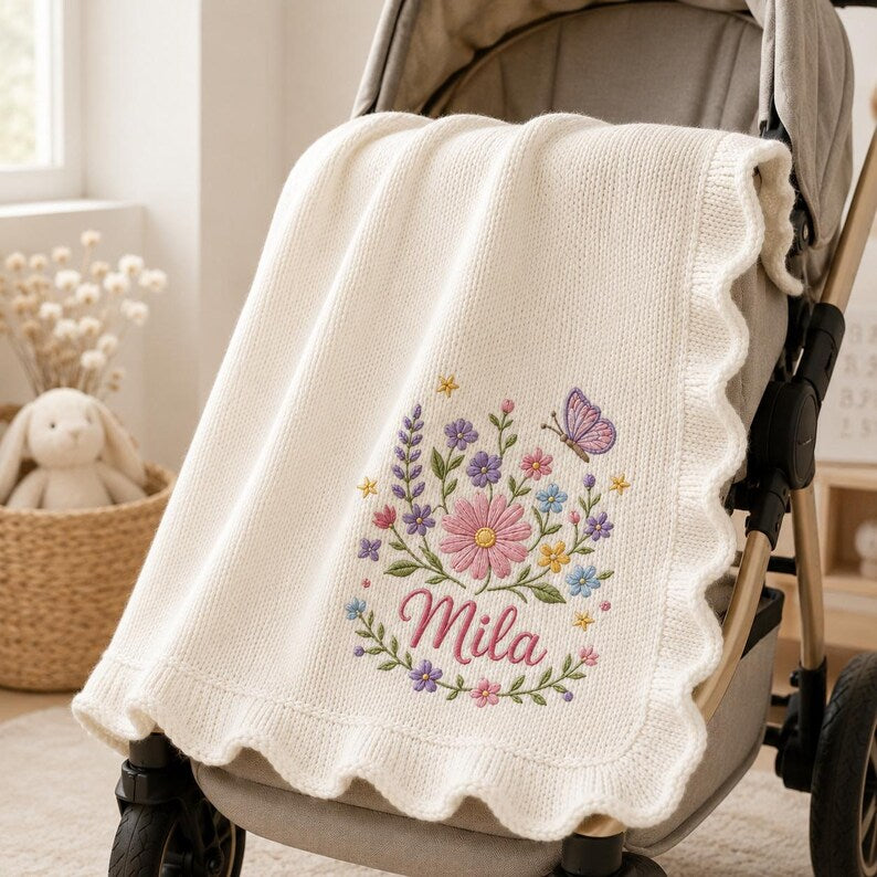 May include: A cream-colored knit blanket with a scalloped edge, featuring an embroidered design of colorful wildflowers, green stems, and a purple butterfly. The name Mila is embroidered in pink script below the floral arrangement.