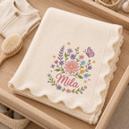 May include: A cream-colored knitted blanket with a scalloped edge, featuring an embroidered floral arrangement of pink, purple, blue, and yellow flowers with a butterfly and the name Mila stitched in pink cursive script in the center.