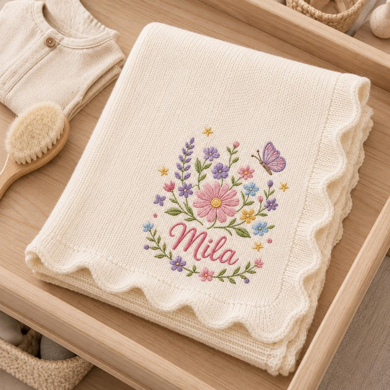 May include: A cream-colored knitted blanket with a scalloped edge, featuring an embroidered floral arrangement of pink, purple, blue, and yellow flowers with a butterfly and the name Mila stitched in pink cursive script in the center.