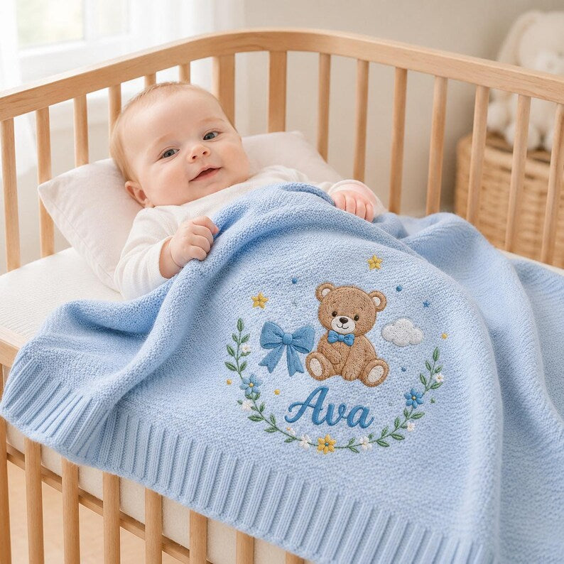 May include: A light blue knitted baby blanket featuring an embroidered design of a brown teddy bear wearing a blue bow tie, surrounded by a floral wreath, a blue ribbon, small yellow stars, and a white cloud, with the name Ava embroidered 