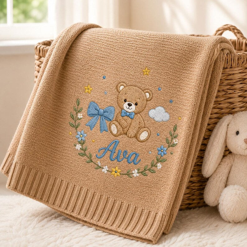 May include: A tan knitted blanket featuring an embroidered design of a teddy bear with a blue bow tie, a blue ribbon, small flowers, stars, and a cloud. The name Ava is embroidered in blue script below the bear, surrounded by a floral wrea