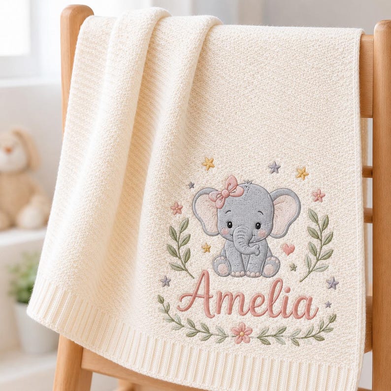 May include: A cream-colored knit blanket featuring an embroidered design of a gray elephant with a pink bow. The elephant is surrounded by green leafy branches, small colorful stars, and flowers. The name Amelia is embroidered in pink scri