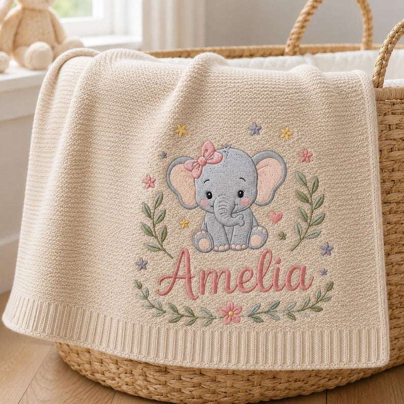 May include: A cream-colored knit baby blanket featuring an embroidered gray elephant with a pink bow, surrounded by green leafy branches, small colorful flowers, and stars. The name Amelia is embroidered in pink script below the elephant.