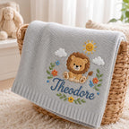 May include: A light gray knitted baby blanket featuring an embroidered design of a brown lion, a yellow sun, white clouds, and colorful flowers. The name Theodore is embroidered in dark blue script below the lion, surrounded by green leave