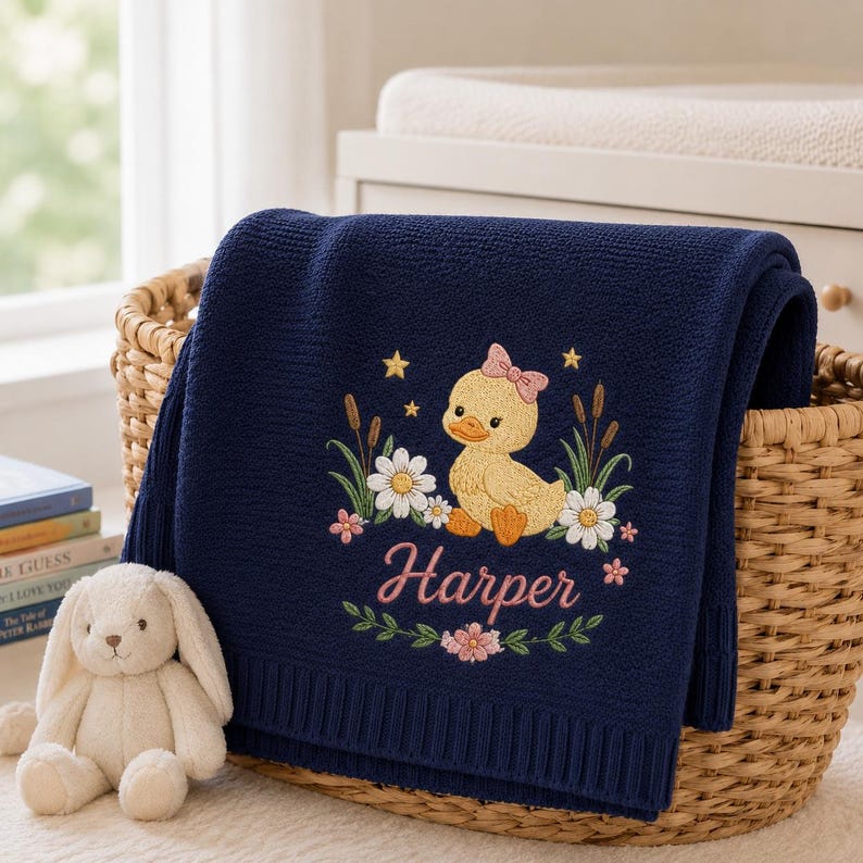 May include: A navy blue knit baby blanket featuring an embroidered design of a yellow duckling with a pink bow, surrounded by white daisies, cattails, and yellow stars. The name Harper is embroidered in pink script below the duckling.