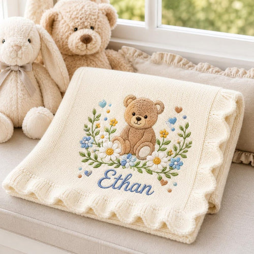 May include: A cream-colored knit baby blanket with a scalloped edge, featuring an embroidered design of a brown teddy bear surrounded by white and blue flowers, green leaves, and small hearts, with the name Ethan stitched in blue script below.