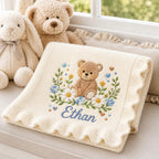 May include: A cream-colored knit baby blanket with a scalloped edge, featuring an embroidered design of a brown teddy bear surrounded by white and blue flowers, green leaves, and small hearts, with the name Ethan stitched in blue script be