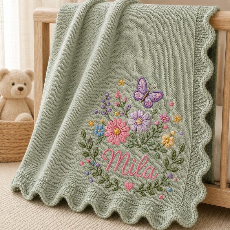 May include: A sage green knitted baby blanket with a scalloped edge. The blanket features an embroidered design of a purple butterfly, various colorful flowers, and green leaves. The name Mila is embroidered in pink script in the center.