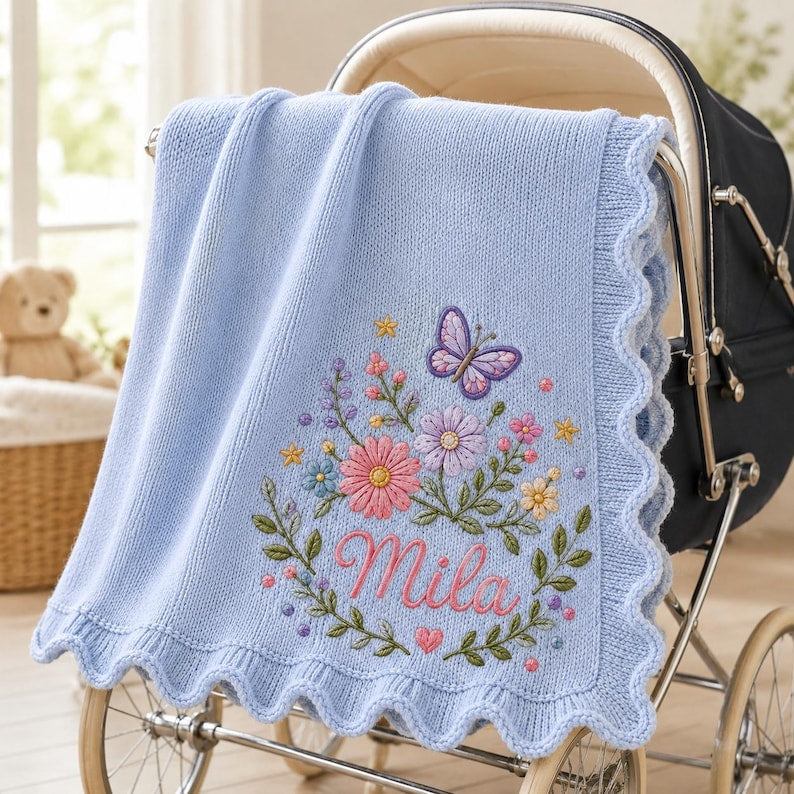 May include: A light blue knitted baby blanket with a scalloped edge, featuring an embroidered design of a purple butterfly, various colorful flowers, green leaves, and the name Mila in pink script lettering, draped over a dark-colored baby