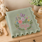 May include: A sage green knitted baby blanket with a scalloped edge, featuring an embroidered design of a purple butterfly, various colorful flowers, and the name Mila in pink script lettering centered within a floral wreath pattern.