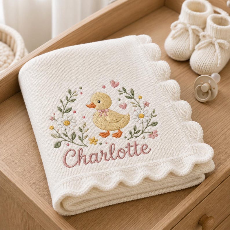 May include: A cream-colored knitted baby blanket with a scalloped edge, featuring an embroidered yellow duckling surrounded by a floral wreath and the name Charlotte in pink script, accompanied by a pair of cream booties and a pacifier.