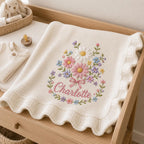 May include: A cream-colored knit baby blanket featuring a ruffled edge and an embroidered floral bouquet in shades of pink, blue, yellow, and white. The name Charlotte is embroidered in pink cursive script below the flowers.