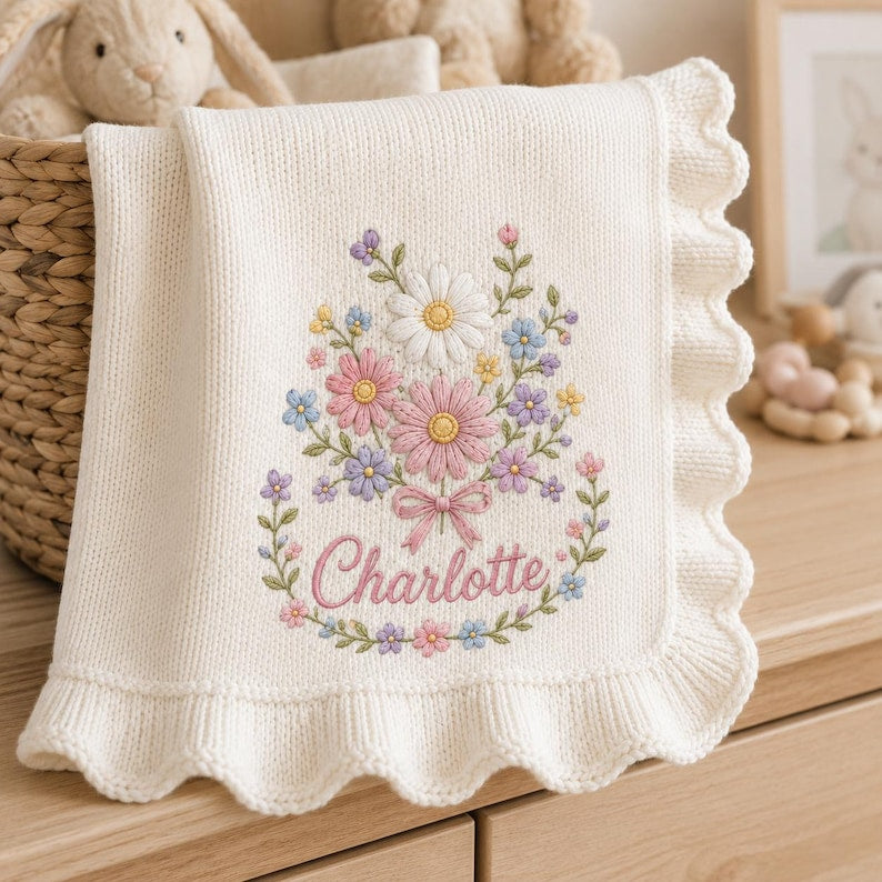 May include: An off-white knitted baby blanket with a scalloped edge, featuring an embroidered floral bouquet in shades of pink, blue, yellow, and white, with the name Charlotte stitched in pink cursive script at the base of the flowers.