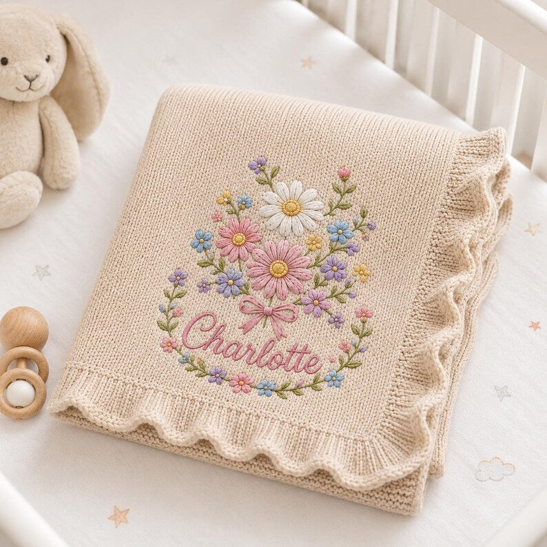 May include: A beige knitted baby blanket with a ruffled edge, featuring an embroidered floral bouquet in shades of pink, white, blue, and yellow, with the name Charlotte stitched in pink cursive script at the center of the flower arrangeme